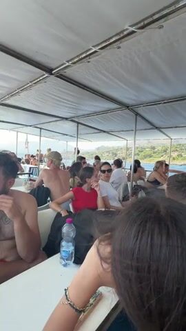 The video shows a group of people on a boat tour. The camera is positioned at the back of the boat, facing forward, with several people seated in the foreground. The boat is moving, and the coastline is visible in the background, with green trees and rocky terrain. The weather appears to be sunny, and the water is blue. The people on the boat are casually dressed, and some are wearing swimwear. They appear to be engaged in conversation or simply enjoying the scenery. Another boat is visible in the distance, also traveling on the water. The atmosphere seems relaxed and vacation-like.