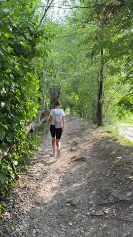 A woman with blonde hair pulled back into a ponytail, wearing a white t-shirt, black shorts, and a small white backpack, walks away from the camera on a dirt path. The path is lined with trees and vegetation on both sides. To the left of the path, a rustic wooden fence runs along the edge of the foliage. The trees are lush and green, creating a canopy overhead, and sunlight filters through the leaves, dappling the path with light and shadow. The ground of the path is covered in dirt, gravel, and some exposed tree roots. The scene suggests a peaceful, natural setting, possibly a park or a hiking trail. The time of day appears to be late morning or early afternoon, given the angle of the sunlight. The weather seems pleasant and sunny. The overall mood is tranquil and serene, evoking a sense of nature and outdoor activity.