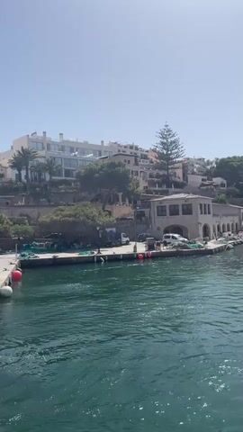 The video shows a wide shot of a body of water, likely a bay or harbor, with a shoreline lined with buildings and vegetation. The water is a vibrant turquoise color and appears somewhat choppy. On the shoreline, there are several buildings of varying sizes and architectural styles. The closest buildings appear to be older, perhaps a commercial or industrial structure with arched openings. Further up the hill, more modern buildings, possibly apartments or hotels, are visible. There are trees and other greenery scattered throughout the scene. Several boats are docked along the shore. The sky is clear and bright blue, suggesting a sunny day. The overall impression is a coastal town or village scene.