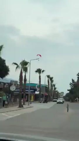 The video shows a street lined with palm trees under a cloudy sky.  A red paraglider is visible in the air between the trees.  On the street, there is pedestrian traffic, including a person walking by a Deniz Market and people riding motorbikes. Several cars are also moving along the street. The video seems to have been taken from inside of a moving vehicle.