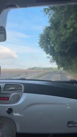 The video shows a view from the interior of a white car, looking out through the windshield. The car is driving on a dirt road. To the left of the road is a field. To the right of the road is a line of trees. The sky is blue with some clouds. The dashboard of the car is visible in the foreground. It is white and has a black trim. There is a phone mounted on the dashboard. The car's radio is on and displaying red text. The video is dated 00:00 to 00:04.