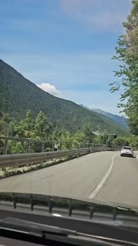 The video shows a view from inside a car driving on a paved road through a mountainous, wooded area. The road curves to the right, with guardrails on the left side of the road. A white car is visible ahead on the right side of the road. The sky is blue with some clouds. Trees and vegetation line the sides of the road, with mountains in the background.
