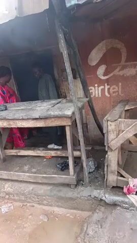 The video shows a group of people standing in front of a small, makeshift structure, possibly a shop or stall. The structure appears to be made of wood and corrugated metal. An Airtel logo is visible on the wall of the structure. A wooden table stands in front of the entrance, and a woman in a red and black patterned dress is standing behind it, interacting with a man. Another man in a white outfit is standing to the left, and a man in a grey outfit is standing to the right. The ground is uneven and appears to be dirt and possibly some rubble. Trash is scattered around the area. The overall scene suggests an informal marketplace or a roadside stall in a somewhat neglected urban environment.