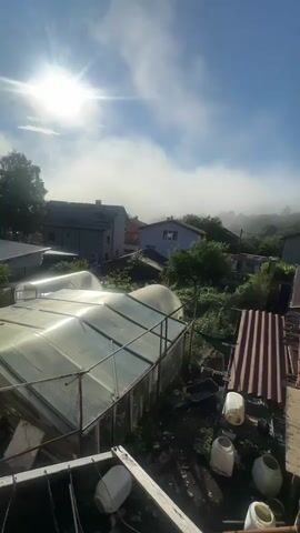 The video pans across a rural landscape under a bright, hazy sun. Several houses with red tile roofs are visible, along with trees and greenery. In the foreground, there is a greenhouse made of clear plastic and metal framing. Other structures, possibly sheds or outbuildings, are also present, one with a corrugated metal roof and another with a weathered wooden frame and a tiled roof. There are several large white plastic containers in the yard. The sky is blue with scattered white clouds and some atmospheric haze or fog in the distance. The overall impression is of a somewhat unkempt but lived-in rural environment.