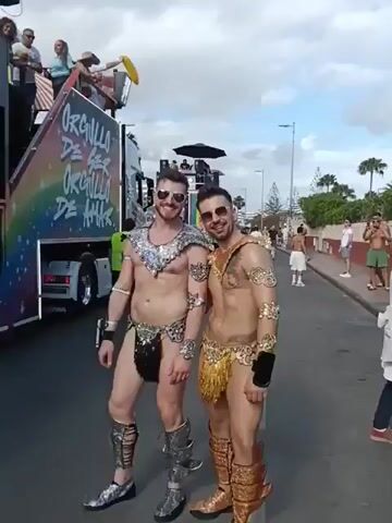Two men in skimpy, glitzy costumes pose on a street beside a parade float. Both men are wearing sunglasses, matching bracelets, and tall decorative boots.  Their costumes consist of a decorative metallic necklace and a small sequined or fringed skirt, leaving their torsos bare. The man on the left has silver accessories while the man on the right wears gold. They turn to face the camera, then turn away and walk down the street.  Behind them is a float with the words "Orgullo de Amor" displayed on the side. People are standing on the float and watching the parade. Other parade watchers are visible along the street.