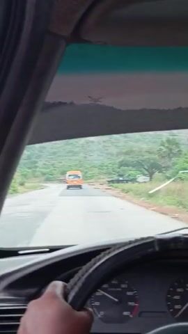 The video is filmed from inside a car, looking forward through the windshield. A dark-skinned hand is visible on the steering wheel in the foreground, with part of the dashboard and speedometer also visible, showing markings for 0, 20, 30, 40, 50, 60, 70, and 80 KM/H. The car is driving on a paved road that appears to be somewhat worn and patchy in places.

Ahead on the road, a small orange van or bus is driving in the same direction. The road curves gently to the left. The scene is set in a hilly and mountainous area, with dense green vegetation, including trees and bushes, covering the slopes. In the distance, on top of one of the hills, a large electrical power line tower (pylon) is clearly visible.

The sky appears overcast and hazy, suggesting a cloudy day. To the right of the road, another vehicle, possibly a dark-colored pickup truck or SUV, is parked or pulled over. The overall setting is a rural or semi-rural landscape. The activity is a car journey through this scenic route, following another vehicle. There is no explicit text visible other than the speedometer markings. The location context provided is Assob Hausa, Nigeria.