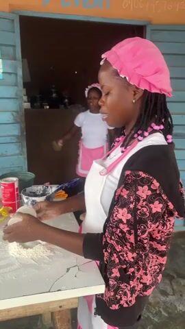 Two young women wearing pink bonnets and aprons are engaged in food preparation at an outdoor table. The woman in the foreground, on the right, is wearing a white apron over a black and pink floral jacket. She is holding a small white container and a white utensil, with flour dusted on the table in front of her. She is looking down at her hands and the container. To her left, a second young woman, also wearing a pink bonnet and white apron over a white shirt, is preparing something from a small packet. She is looking at the packet in her hands. On the table between them are a rolling pin, a can of soda, and a bowl containing a blue substance. The background appears to be an open-air structure with blue wooden siding and an orange awning. A sign with blue text is visible on the siding.
