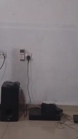 The video shows a wall with electrical outlets and several audio devices on the floor. On the left is a tall, black speaker. To its right is a square, black subwoofer with a circular opening. In front of the subwoofer is a flat, black electronic device, possibly a DVD player or Blu-ray player, with wires connected to it. Another black electronic device is stacked on top of it. There are also a few CDs stacked on top of the subwoofer. The wall is painted white and has two electrical outlets visible. The floor is tiled. The video pans slowly from left to right, showing the arrangement of the devices.