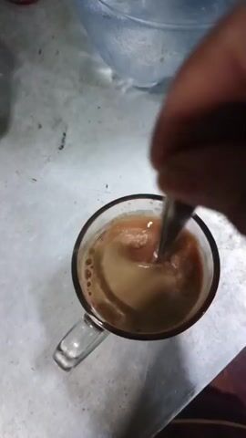 A hand holds a metal spoon, stirring a light brown liquid in a clear glass mug. The liquid is in motion, creating ripples and swirls. The mug has a handle and sits on a speckled white surface. In the background, a blurry bowl is visible.