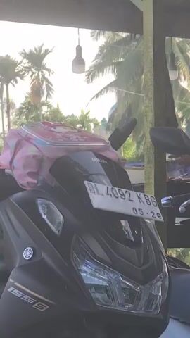 The video shows a roadside stand selling coconuts, likely in a tropical or subtropical region. Several motorbikes are parked outside the stand.  Two young men, one wearing a cap, sit on one of the motorbikes, facing away from the camera. A woman wearing a hijab tends to the stand, which is stacked high with green coconuts and some ripe oranges. The stand has a thatched roof and green wooden supports. Beyond the stand, there are glimpses of trees and buildings in the background, suggesting the location is an inhabited area near nature.