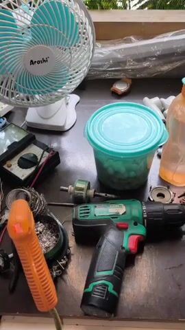 The video shows a cluttered workbench with various electronic components and tools. In the foreground, an orange soldering gun is visible, connected to wires. To its left, a spool of solder and a multimeter are present. A green and black cordless drill is positioned in the center, with a small motor and other parts scattered around it. In the background, a white desk fan with a blue grill is on, and various wires, capacitors, and a plastic container filled with small objects are also visible. The overall impression is one of ongoing repair or assembly work.