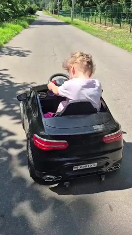 A young child with blonde hair is sitting in the driver's seat of a black toy car, steering it forward on a paved road. The child is wearing a white t-shirt. The toy car appears to be a replica of a Mercedes-Benz, with realistic details like headlights, taillights, and exhaust pipes. The car has a license plate that reads "X001XX." The road is lined with trees and grass, and a white fence is visible on the right side of the road. The sky is bright, suggesting it is daytime. The child is facing away from the camera, focused on driving.