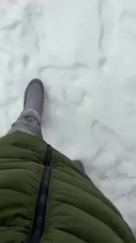 The video shows a person standing in snow. The individual is wearing a puffy green coat and gray shoes. They hold up a white paper coffee cup with a lid that has a small brown stain around the opening. The cup is labeled "COFFEEIN" in black lettering. The environment is snowy, with a tree visible in the background.