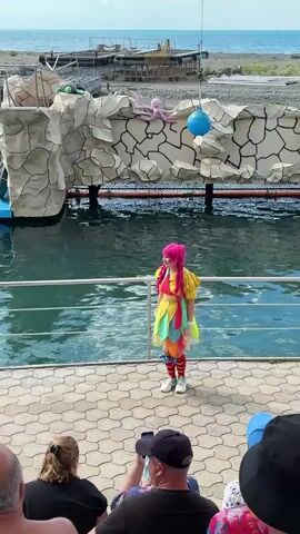 A video shows a costumed female performer with bright pink hair standing on a stone-paved stage in front of a body of water. She wears a yellow shirt, a colorful skirt with feather-like segments, striped red and white leggings, and white shoes and gloves. Behind her, a rocky-looking backdrop features a pink octopus and a blue sphere. The water beyond the stage appears green, and in the distance, a blue sky meets the horizon above what looks like a distant shoreline. Several spectators are visible in the foreground, seated with their backs to the camera, observing the performance.