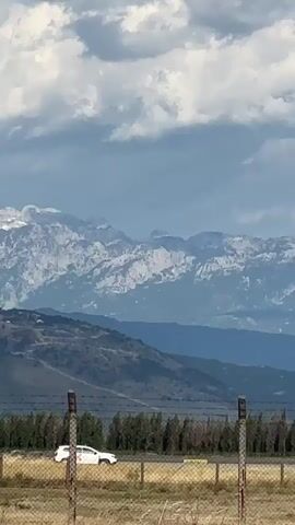 The video captures a scenic view of mountains with snow-capped peaks under a cloudy sky. In the foreground, a fence with barbed wire encloses a dry, grassy field. A white car with a red and white logo on its side is visible on a road in the middle ground. A dark-colored helicopter flies horizontally across the frame from left to right, positioned between the car and the mountain range. The helicopter appears to be in mid-flight, with its rotor blades spinning. The overall environment suggests a rural or semi-rural location, possibly near an airport or an open area suitable for helicopter operations.