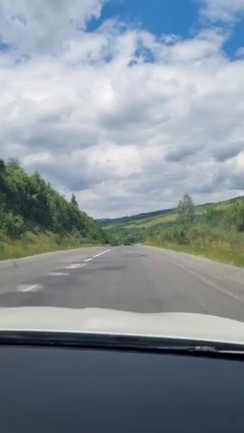 The video shows a view from inside a car driving on a road. The road has some potholes and is bordered by green trees and hills on both sides. The sky is blue with scattered clouds. There are a few cars visible in the distance on the road. The video appears to be taken during the daytime.