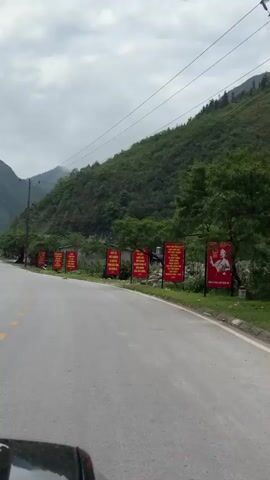 The video shows a two-lane paved road winding through a mountainous, heavily forested region. The weather appears overcast. Along the roadside are several red signs with yellow text. Two motorcyclists are seen traveling along the road, away from the camera. A large truck is parked further up the road. Utility poles and power lines run alongside the road. The vegetation is lush and green.