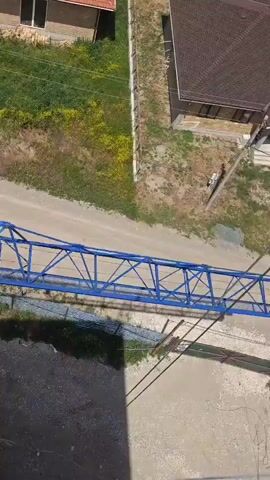The video shows an aerial view of a construction site. A large blue crane boom is being assembled or disassembled. The crane boom is extended horizontally and appears to be leaning against a tall, dark vertical structure, possibly a chimney or a temporary tower. The boom is made of a metal lattice structure. In the background, there is a dirt road with a few parked cars. There are also some buildings and grassy areas. The lighting suggests it is daytime with a bright sky and some shadows. There are no people visible in the footage.