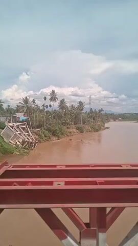 A bridge railing, painted red, is in the foreground. Beyond the railing, a muddy brown river flows under a cloudy sky. On the riverbank, palm trees and dense vegetation are visible. In the distance, mountains can be seen. An Indonesian flag, red and white, is flying from a pole on the bridge.