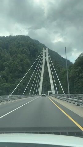 This video shows a view from a car driving towards a modern cable-stayed bridge. The bridge has two tall, triangular concrete towers connected at the top, from which numerous white cables extend downwards, supporting the bridge deck. The bridge spans across a valley with steep, densely forested mountains on either side. The road has multiple lanes, marked with white and yellow lines. There is a tunnel visible at the end of the bridge, and another car is driving in the distance. The sky is overcast with thick, dark clouds. The camera is positioned inside the vehicle, looking forward through the windshield.