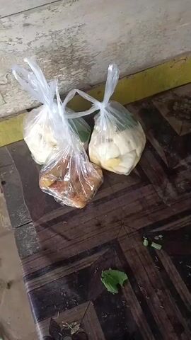The video shows three plastic bags filled with food, arranged on a wooden table. Two of the bags contain pale, possibly fruit or vegetable, items, while the third bag holds brown, cooked food. Several green leaves and small chopped green pieces are scattered on the table. Further down the table, there are two cups with lids, one containing a dark liquid and the other a lighter colored liquid, and a lighter. The background shows a textured wall and a wooden surface.