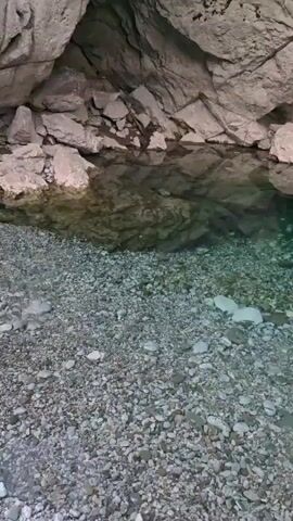 The video shows a view of a clear shallow body of water with a rocky shore and a cave entrance in the background. The water is transparent, allowing visibility of the pebbles on the bottom. The cave walls are made of rough, light-colored rock. The lighting suggests an overcast day or the shade of the cave.