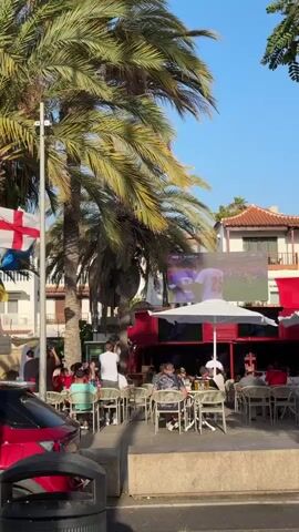 A group of people are seated at tables outside a building with palm trees and a large screen showing a soccer game. Some people are standing and cheering, others are seated and watching the game. The screen displays players in white and black uniforms. The environment appears to be a sunny outdoor cafe.