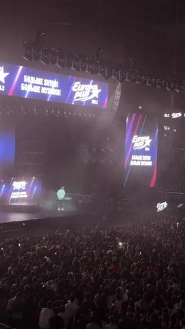 A concert is taking place indoors in a large arena. The stage is brightly lit with screens displaying the Europa Plus logo and the text "БОЛЬШЕ ХИТОВ! БОЛЬШЕ МУЗЫКИ!" (More hits! More music!). A lone figure, likely a DJ or performer, stands at a console on the stage, silhouetted against the lights. Smoke or fog machines are active, creating a hazy atmosphere. A large crowd of spectators fills the arena, many holding up their phones, suggesting they are recording or taking pictures. The lighting is dramatic, with spotlights and flashing effects. The overall environment suggests a high-energy event.