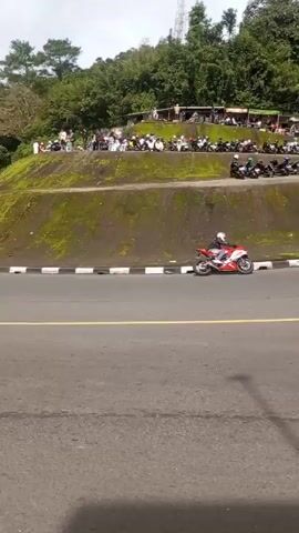 The video shows a busy two-lane road with a line of spectators and motorcycles parked along the shoulder.  The road curves uphill, with a backdrop of lush green trees covering the hillside. Cars and motorcycles travel along the road. The video ends at an intersection with a petrol station visible.