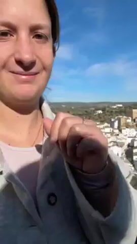 A woman stands outdoors on a sunny day, holding a smartphone and recording herself. She is fair-skinned, has brown hair pulled back from her face, and is wearing a light gray jacket over a pink top.  In the background is a scenic view of a village with white buildings and a tower, set amidst rolling hills and trees under a bright blue sky. A metal railing is visible in the foreground, suggesting she is on a balcony or overlook. The camera is handheld and slightly shaky. She glances at the camera and smiles slightly. A man's voice, likely in the background, is audible.