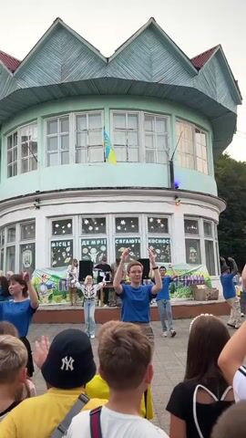 The video captures an outdoor event in front of a multi-story building with a distinctive turquoise roof. A group of people, predominantly young adults and children, are gathered in a courtyard area. A stage is set up, featuring individuals performing or DJing, with a banner behind them that appears to have text in Ukrainian. Several young people in blue shirts are actively dancing in unison in the foreground, facing away from the camera. Other spectators are visible, some recording the event on their phones. The building itself has large windows, and Ukrainian flags are displayed. The atmosphere suggests a public gathering or performance.