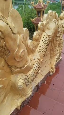 A close-up shot focuses on the scales and golden features of a dragon statue. The camera pans right, revealing more of the dragon's body, including its claws. Further panning shows the dragon's head, characterized by its open mouth, sharp teeth, and large black eye. The statue is adorned with ornate, cloud-like carvings and sits atop a red staircase. In the background, lush green foliage and distant buildings are visible under an overcast sky.