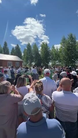 A video captures an outdoor gathering under a bright, sunny sky with scattered clouds and green trees in the background. A small wooden building with a red roof is visible on the left. A crowd of people, seen from behind, are facing a central area where a group of individuals, some in light green outfits, appear to be performing or dancing. Several people in the foreground are holding up their phones, seemingly recording the event. The atmosphere appears festive and engaged, with a sense of community participation. The overall setting suggests a cultural event or celebration taking place in a rural or park-like environment.