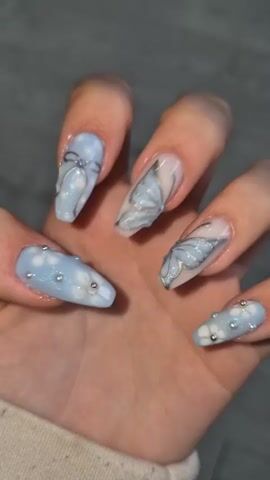A close-up shot features a well-groomed human hand, likely female, showcasing an elaborate set of decorated fingernails. The nails are long and shaped in a coffin or ballerina style, predominantly adorned with light blue, white, and silver accents, with some clear or nude base visible on specific nails.

The index finger features a light blue base with a detailed silver bow design, accented by a small gem at its center, against a subtle white, cloud-like pattern. The middle and ring fingers display prominent, intricate butterfly motifs. These butterflies are rendered in light blue with elegant silver outlines and internal details, enhanced by a touch of glitter, set against a clear or nude nail base.

The thumb, partially visible, and the pinky finger, along with another finger on the lower left (appearing to be the middle finger due to its length and decoration matching the other blue nails), are painted with a light blue base. These nails are covered with small white flower patterns and dotted with tiny silver beads, adding texture and sparkle.

The hand itself has a fair skin tone. The background is a soft, blurry grey or light blue, suggesting an indoor setting. The lighting appears to be artificial, highlighting the glossy finish and intricate details of the nail art. No specific activity is occurring other than presenting the nails. No text is visible in the image.