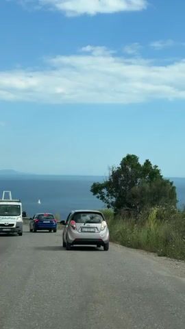 A car is driving on a road alongside the sea. There are other cars driving in front of it, and a metal guardrail is visible on the right side of the road. The sea is deep blue, and a small sailboat can be seen in the distance. The sky is blue with some scattered white clouds. Some roadside vegetation is also visible.