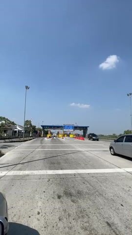 The video shows a highway toll booth on a clear day. There are multiple lanes leading up to the toll plaza, with some cars already present. The toll plaza itself has several booths with barriers and signs. The surrounding environment appears to be somewhat rural or suburban, with some buildings visible in the distance and trees. The sky is blue with a few scattered clouds. The camera appears to be mounted on a vehicle, as evidenced by the perspective and slight motion.