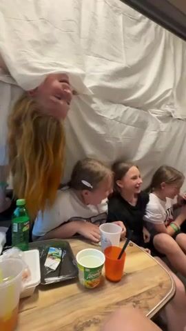 A group of young girls are sitting inside a cramped, dimly lit space, possibly a tent or RV. They are gathered around a wooden table filled with snacks and drinks. The walls of the space are covered with a white, draped material, creating a makeshift enclosure. The girls appear to be engaged in conversation and are visibly animated, with some of them laughing and gesturing. The overall atmosphere suggests a playful and intimate gathering.