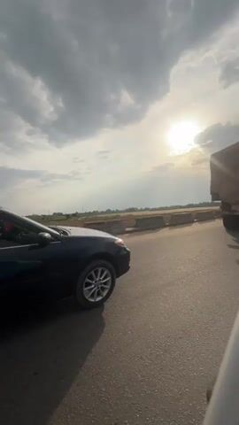 This is a vertical, low-angle, medium shot video clip. The video focuses on a dark blue sedan parked on a paved road. The sun is visible in the upper right quadrant of the frame, partially obscured by clouds, casting a bright glow. The sky is filled with a mixture of light and dark clouds. The car is positioned to the left of the frame, with its side facing the camera. The front of the car is facing right. The driver's side door is open, and a person is visible in the driver's seat, wearing a white shirt. The car's interior is dark. The road surface appears to be asphalt. In the background, there is a road divider, a truck, and some vegetation. The overall lighting suggests it is either sunrise or sunset.