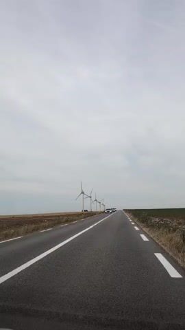 The video shows a view from a car driving down a road. Wind turbines are visible on the left side of the road, and fields are on either side. Several white cars are driving ahead on the same road. The sky is overcast.