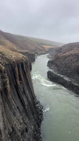 The video captures a wide, scenic view of a deep river canyon, likely in Iceland, characterized by striking geological formations. A substantial, fast-flowing river, with a greenish hue and visible white water rapids, carves its way through the landscape.

The most prominent feature is the left canyon wall, which displays a dramatic, sheer cliff face composed of dark, well-defined hexagonal basalt columns, extending vertically from the water's edge. Similar, though less extensive, basalt columns are also visible along the right bank of the river. The surrounding terrain consists of undulating hills covered in sparse, brownish vegetation, indicative of a rugged, possibly volcanic environment.

The sky is entirely overcast, casting a soft, diffused light over the scene, suggesting a cloudy day. As the video progresses, the camera slowly pans from left to right, gradually revealing more of the expansive canyon. Two small human figures can be seen walking along a path on the upper rim of the right canyon bank; one of them is notably wearing a red jacket. The panning motion emphasizes the vastness and natural grandeur of the canyon and the powerful river flowing through it. No text is visible in the video.