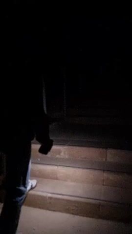 The video begins in near darkness, showing the lower half of a person walking up a set of stone steps. The person is wearing jeans and boots and appears to be holding something in their hand. The steps are dimly lit, and a metal railing is visible in the background. The video then cuts to a dark scene that reveals a view of a town or city at night, with lights from buildings and streets twinkling in the distance. The focus is soft, making the lights appear somewhat blurry.