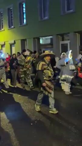 A small parade, possibly celebrating carnival, travels down a narrow street at dusk. Costumed individuals play instruments including a tuba, French horn, and clarinet, while walking amid others in costume. The costumes include animals, a mariachi musician, and a person in orange. Buildings painted green line the street. The parade moves from left to right in the frame, with spectators visible on the right side of the road.