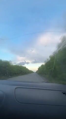 A POV shot from inside a car driving down a road lined with trees. The sky is blue with clouds, and the sun is setting in the distance. The windshield is dirty and has a crack. The car's dashboard is visible in the foreground.