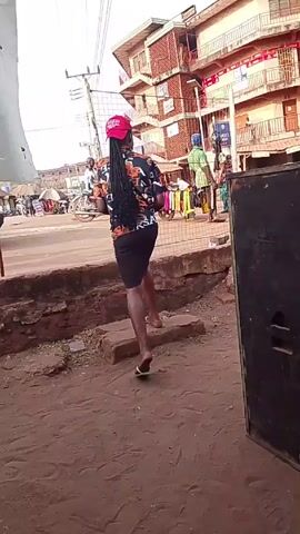 The video shows a street scene with people walking on and around a low brick ledge. A person with long dark hair wearing a red baseball cap, a colorful shirt, and a short dark skirt walks along the ledge, performing a high leg kick. Another person walks along the ledge behind her, carrying a bag. A cart of colorful clothing is visible in the background, and other people are seen walking around. The background also includes buildings with multiple stories, and power lines overhead. A large, rectangular speaker sits in the foreground on the right side of the frame. The ground is a reddish-brown color, suggesting a dirt or clay surface.