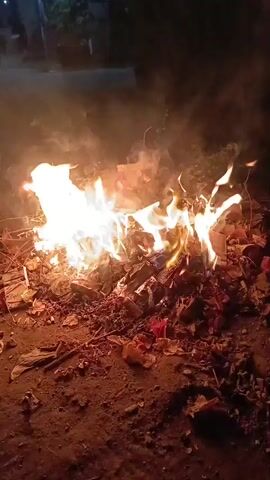 The video shows a pile of burning trash at night. Flames are visible, and smoke rises from the fire. The trash appears to consist of dried leaves, twigs, and possibly paper products. The ground around the fire is dirt and debris. The background is dark, suggesting it is nighttime. There are no people or animals visible in the video.