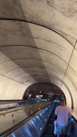 The video shows a view from behind a person and a child standing on an escalator in a subway station. The escalator is moving downwards, indicated by the person's position and the perspective of the scene. The escalator has chrome-plated handrails that reflect the blue lighting from below. The ceiling of the station is made of concrete and is arched, with shadows cast from overhead lights. There are other escalators visible, also moving downwards. The overall environment appears to be a modern subway system. The people are facing away from the camera and towards the bottom of the escalator.