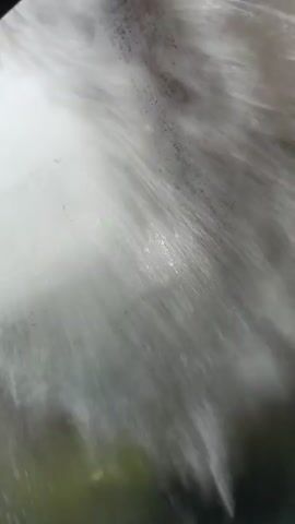 A close-up, low-angle shot captures water being sprayed onto a surface. The water creates frothy, white bubbles and streaks as it cascades downwards, obscuring much of the underlying material. The surface appears to be a metallic or reflective material, possibly the floor of a car wash, with hints of green and yellow visible through the water in the lower portion of the frame. The spray appears to be forceful, creating a dynamic and somewhat abstract visual effect.