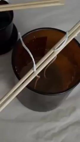 The video shows two amber-colored glass candle containers, one larger and one smaller. Each container holds candle wax and a white wick. Two wooden chopsticks lay across the top of each container, suspending the wicks in the center. The containers sit on a white, textured surface.