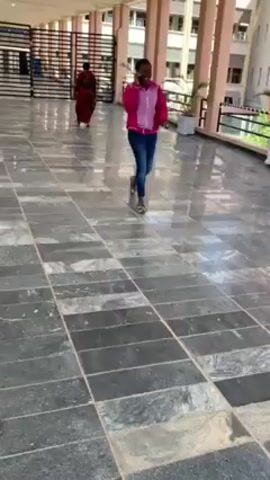 A woman is walking and talking on her phone on a tiled walkway. She is wearing a pink and light pink jacket, jeans, and slides. She walks toward several people standing by a railing and some video equipment. The walkway is covered in dark grey tiles, and there are pillars supporting a roof above. Beyond the railing is a view of a desert-like area.