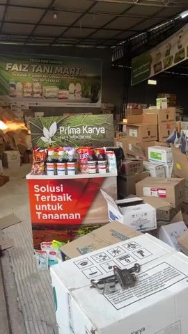 The video pans across a cluttered storage area, likely a warehouse or a shop backroom, filled with numerous cardboard boxes and bags of supplies. The boxes are stacked haphazardly, some open and others sealed, with various labels and packaging visible. In the foreground, a large white box with a padlock is prominent. To the left, a display stand features bottles of what appear to be agricultural products, possibly pesticides or fertilizers, with prominent branding. In the background, banners and signage suggest the establishment is a farming supply store, possibly named "AIZ TANI MART." The overall impression is one of disarray and a large volume of stored goods. There are no people visible in the frame.